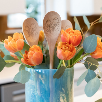 Wooden spoons with floral designs in a blue vase with orange tulips and green leaves.